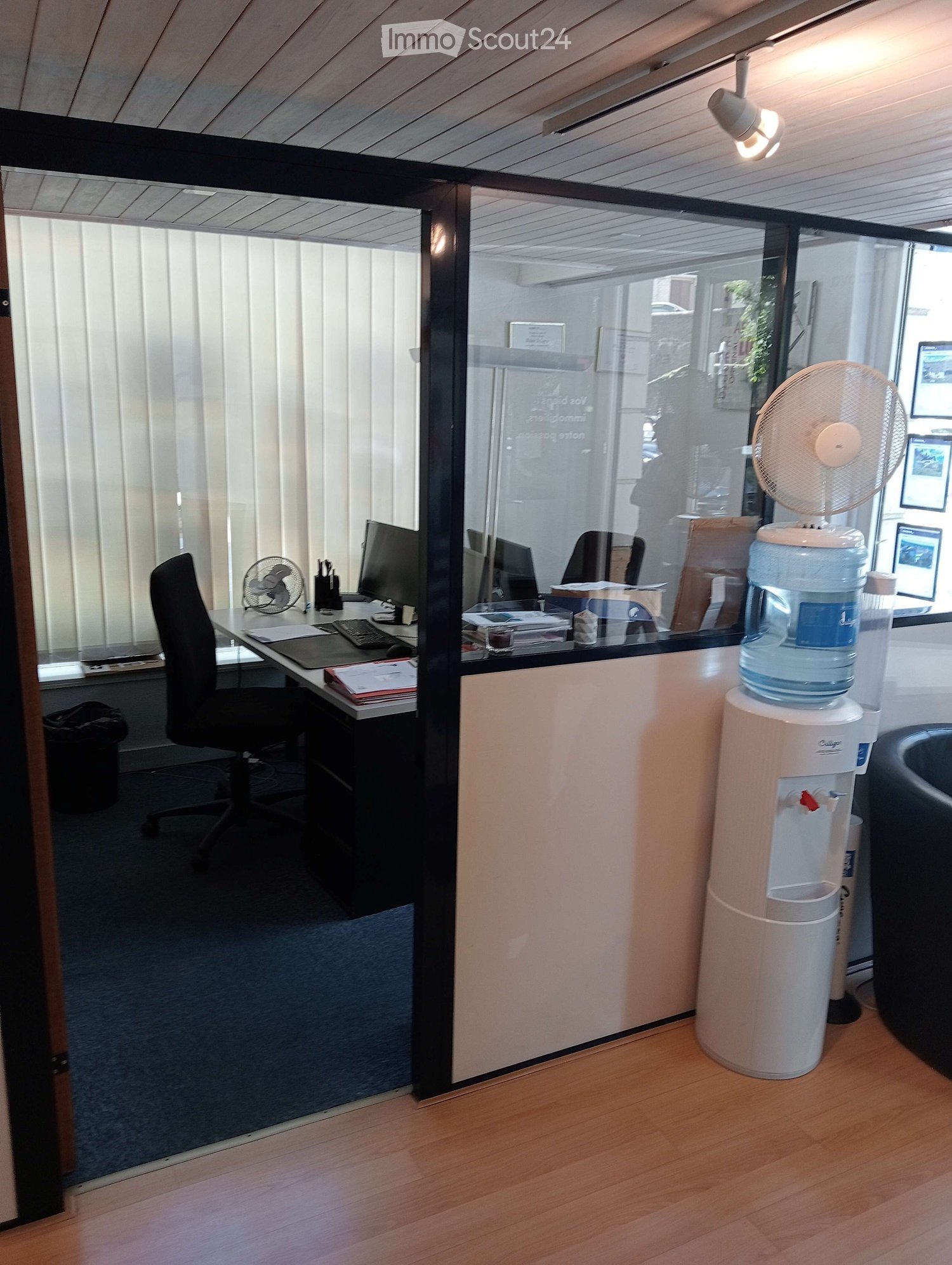 Office space with a desk, office chair, computer monitor, keyboard, water cooler, and fan. The room has glass partitions and a wooden floor.