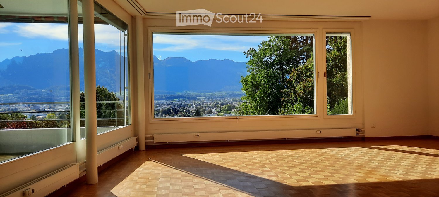 Empty room with wooden flooring, large windows offering a scenic mountain view, and a balcony area visible on the left.