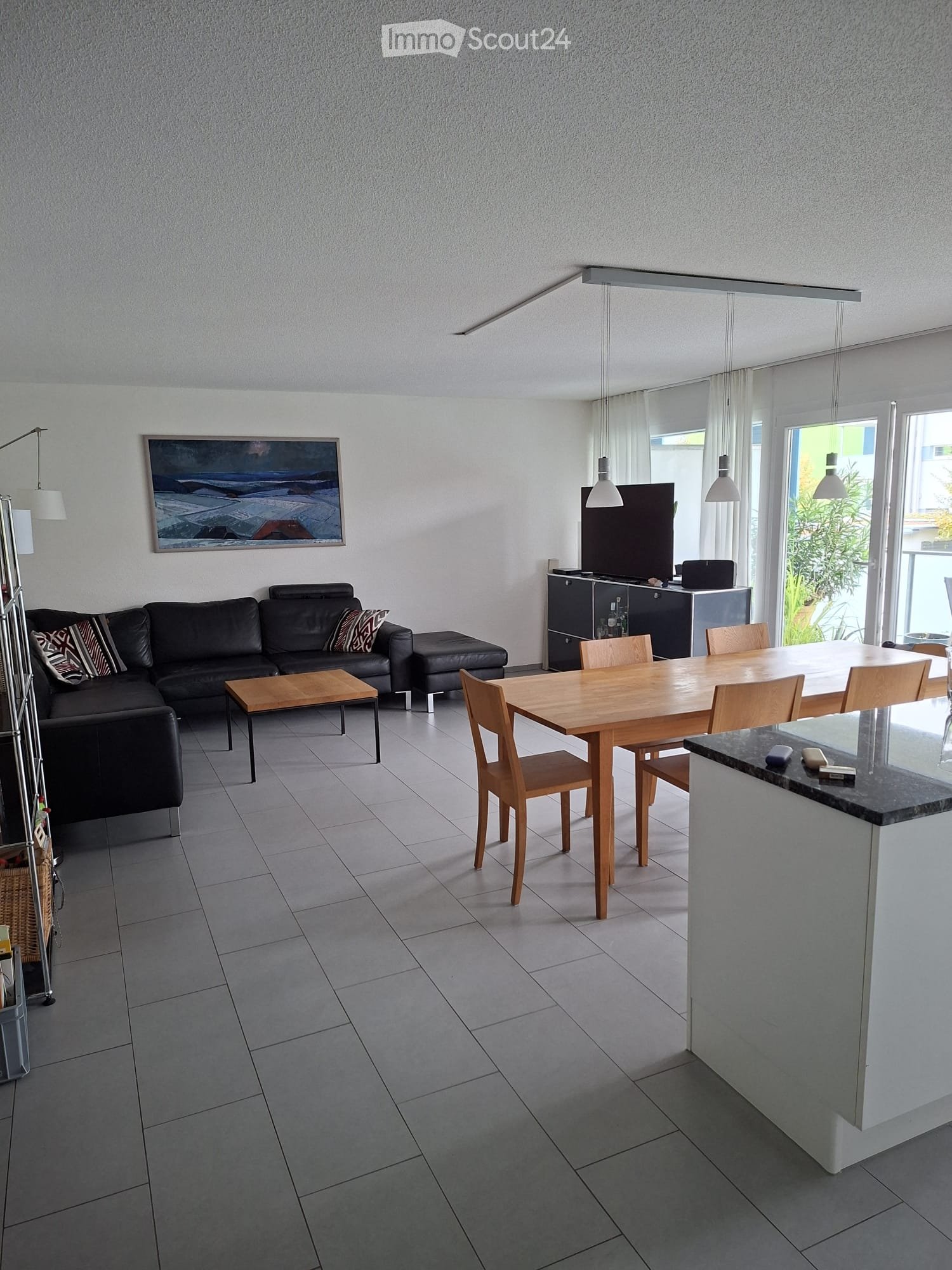 Open-plan living and dining area with a grey tiled floor, a black leather sofa, a wooden coffee table, a dining table with chairs, and a TV on a stand.