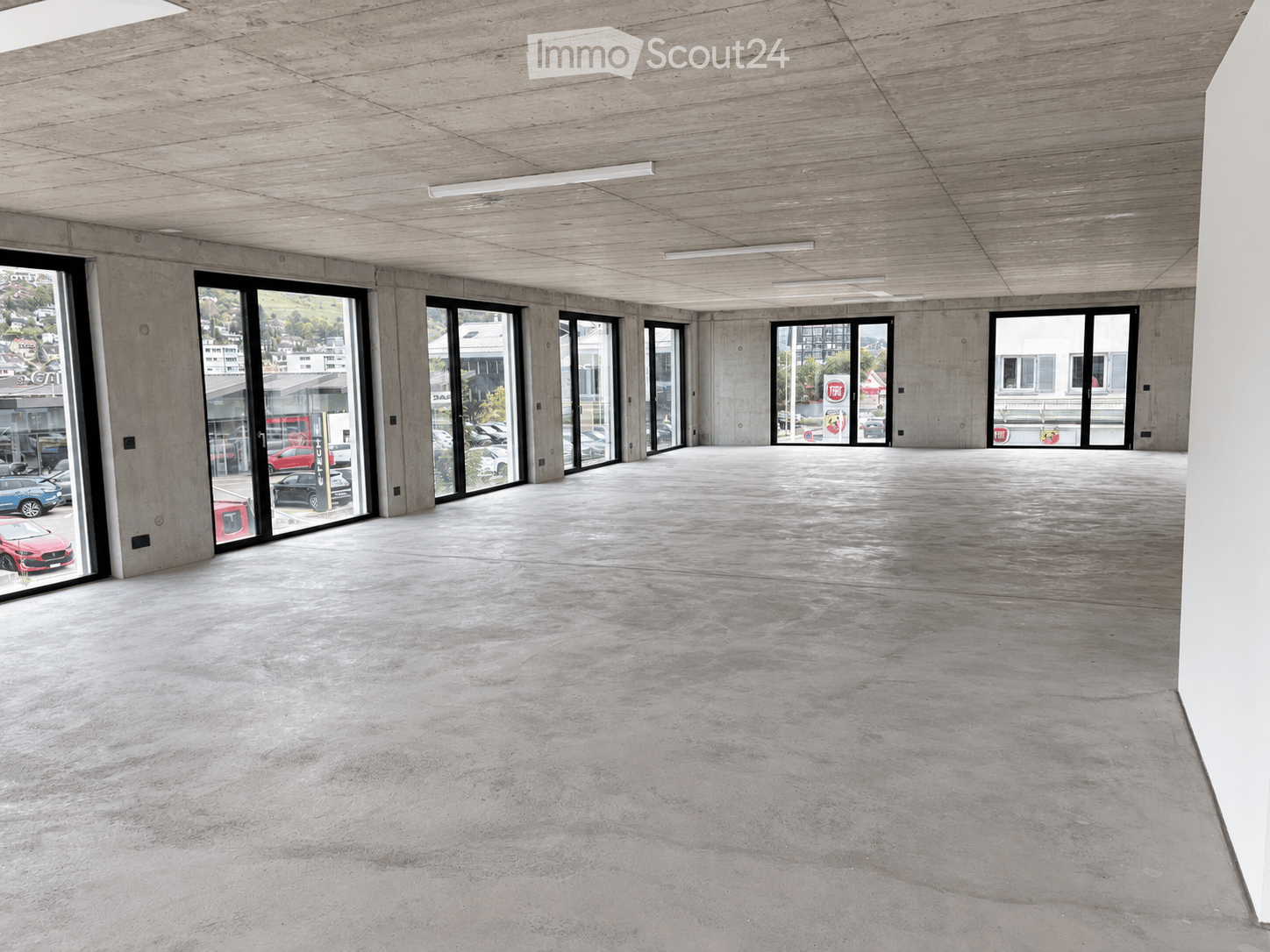 Empty room with concrete floors and walls, large glass windows, and exposed ceiling beams.