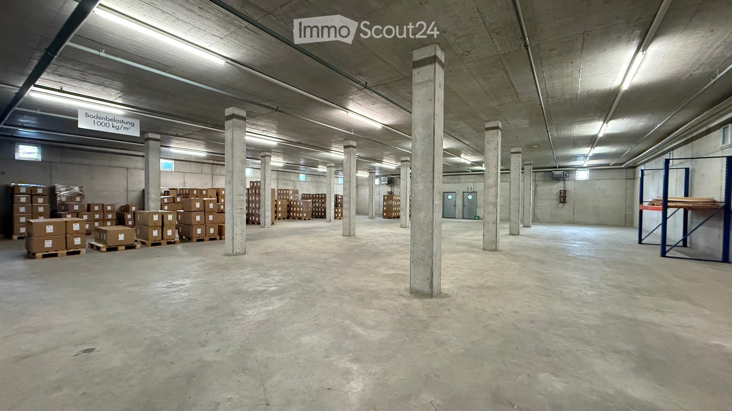 Empty warehouse space with concrete floors and pillars, storage boxes, and a sign indicating floor load capacity.