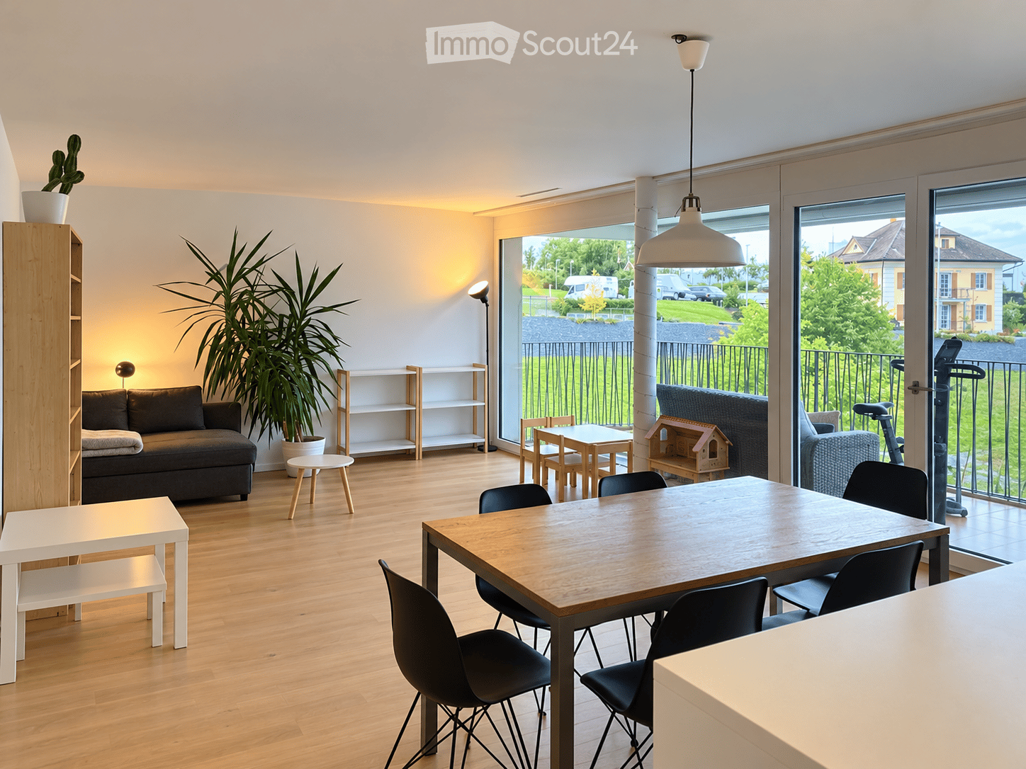 Bright living and dining area with wooden flooring, large windows, a sofa, dining table with chairs, shelves, and potted plants.