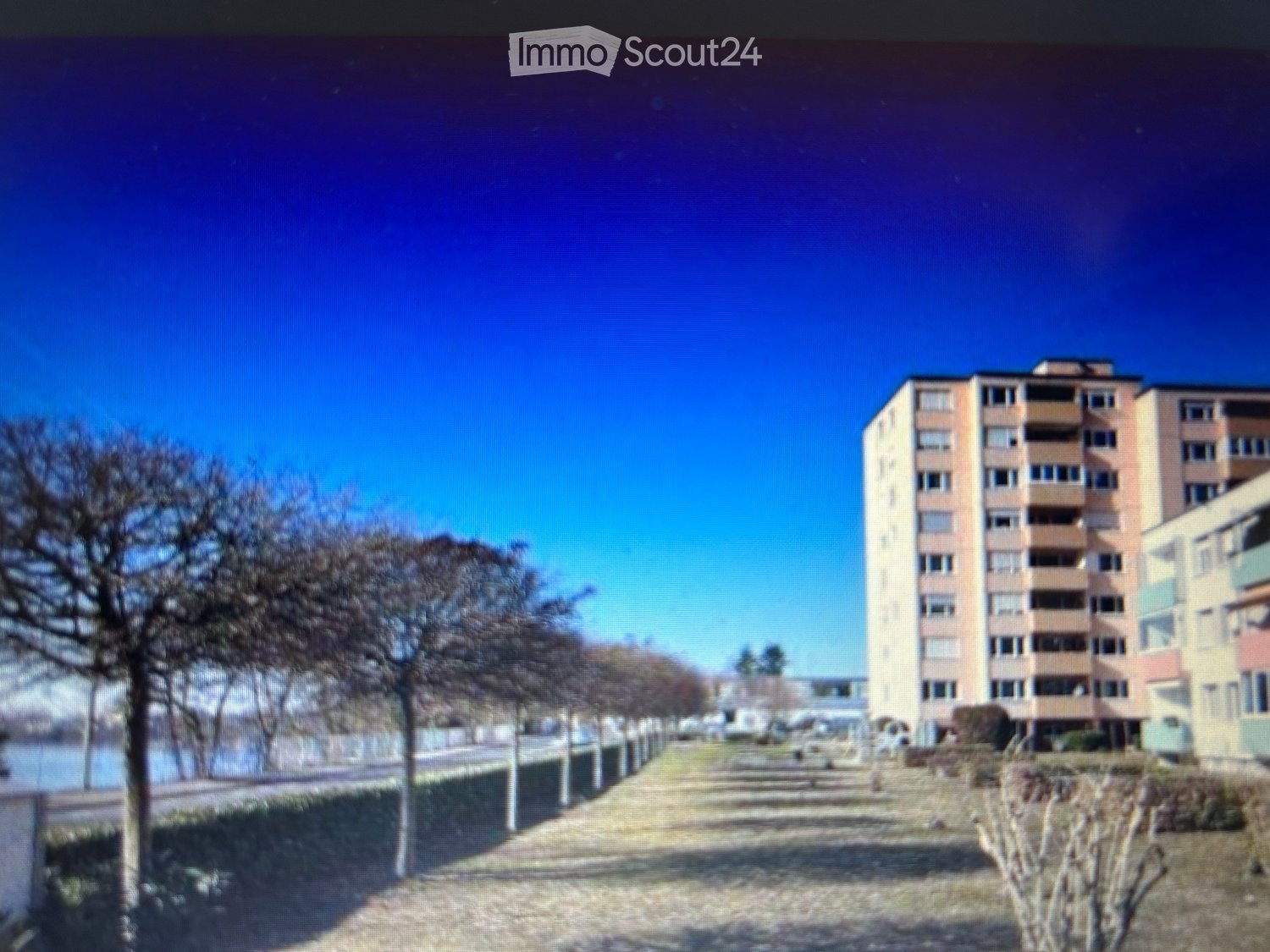 Multi-story residential building with balconies and windows, situated near a waterfront with trees and a paved pathway.