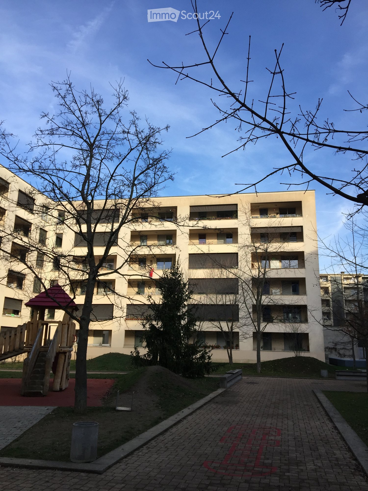 Modern multi-story residential building with balconies and large windows, surrounded by trees and a small playground area.