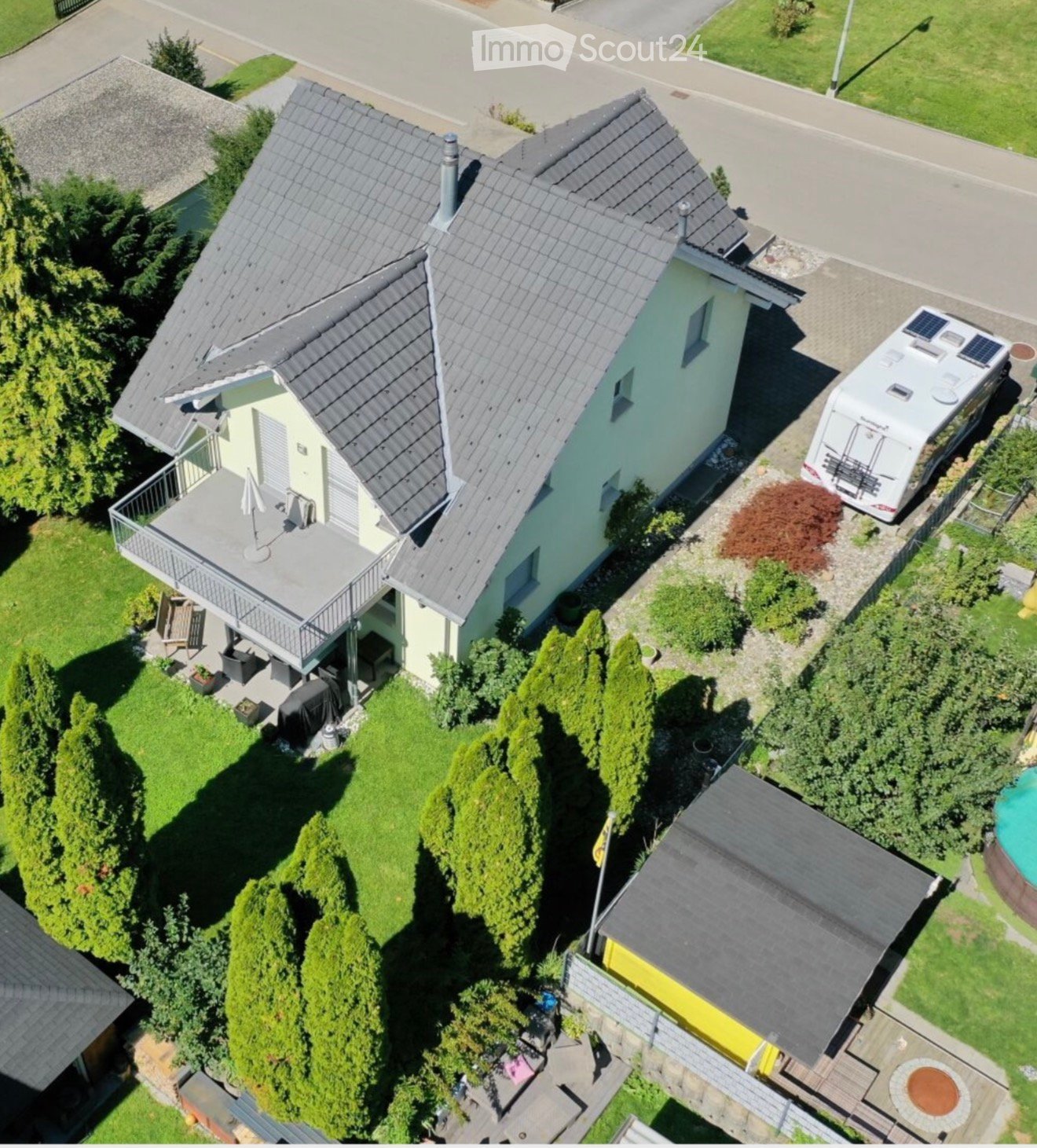 A two-story house with a grey roof and light yellow walls, featuring a balcony, garden, and a camper van parked nearby.