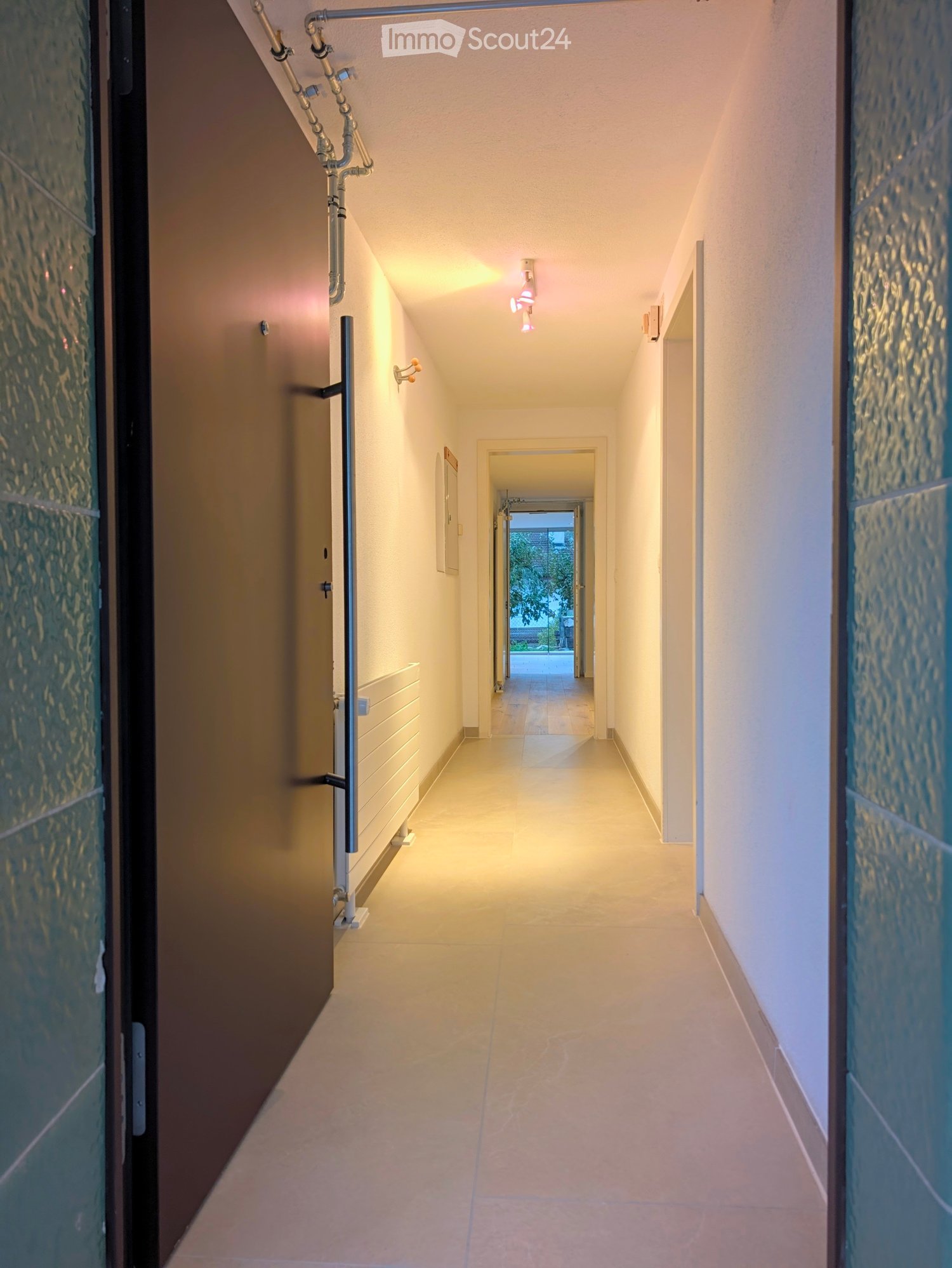 Clean, well-lit hallway with beige tiled flooring, white walls, and modern lighting fixtures. Features a door on the left and a glimpse of an outdoor area at the end.