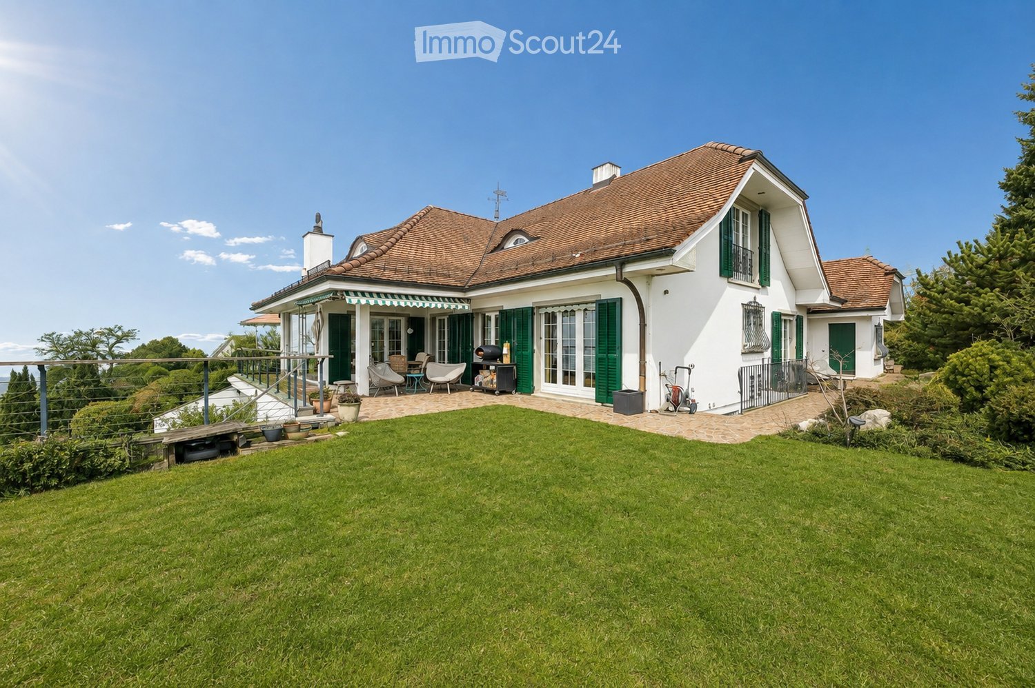 A two-story house with a red-tiled roof, white exterior, and green shutters. The house features a covered porch with outdoor seating, a well-maintained lawn, and a patio area with a grill and outdoor furniture.