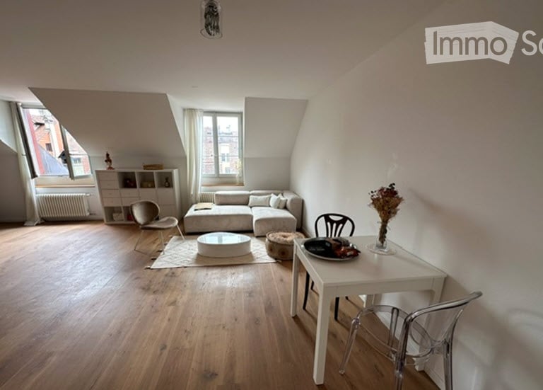 Bright, open living space with wooden flooring, white walls, and large windows. Features a grey sectional sofa, white coffee table, dining table with chairs, and a small bookshelf.