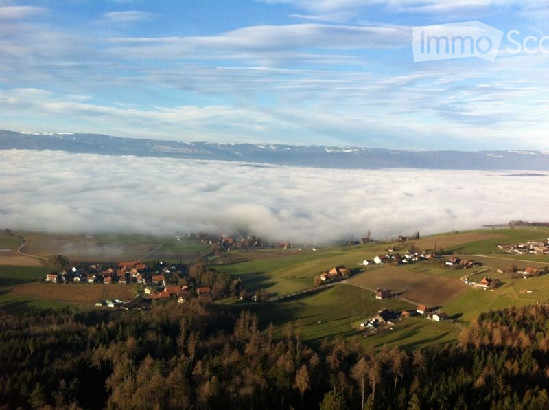 Aerial view of a rural village nestled in a valley with scattered houses, fields, and a forested area, surrounded by misty mountains under a clear blue sky.