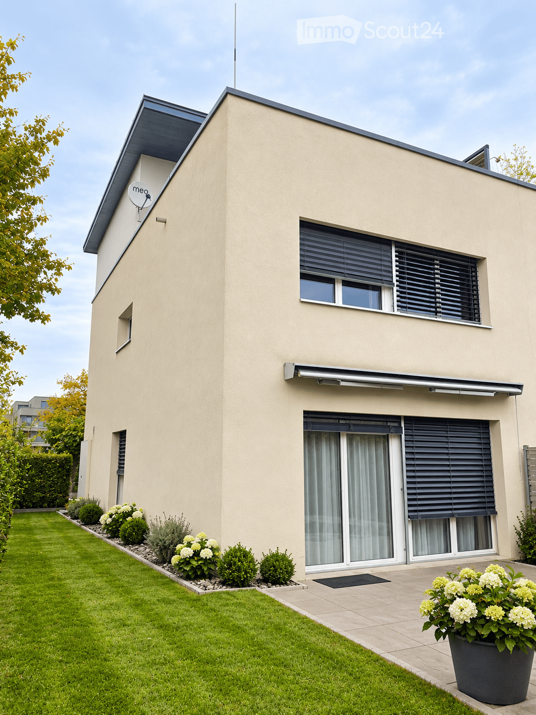 Modern two-story house with beige exterior, large windows, sliding glass doors, and a well-maintained garden with shrubs and flowers.