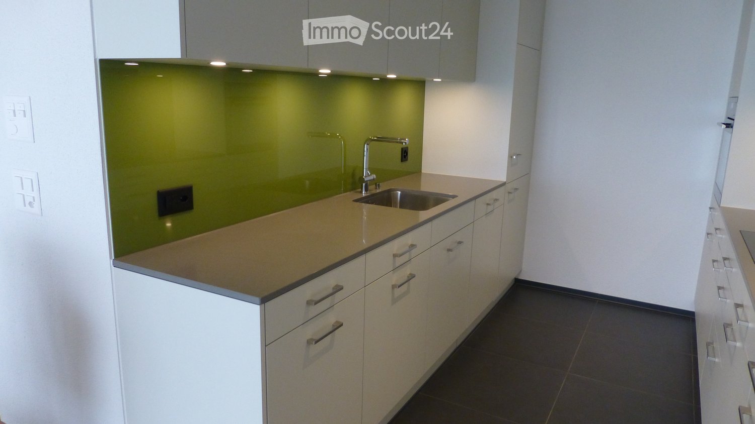 Modern kitchen with white cabinets, beige countertop, and a green backsplash. Features a stainless steel sink and faucet.