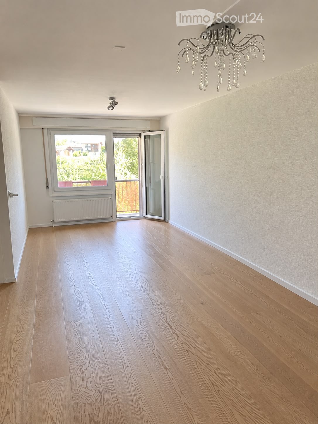 Empty room with light wooden flooring, white walls, a chandelier, and a large window with sliding doors leading to a balcony.