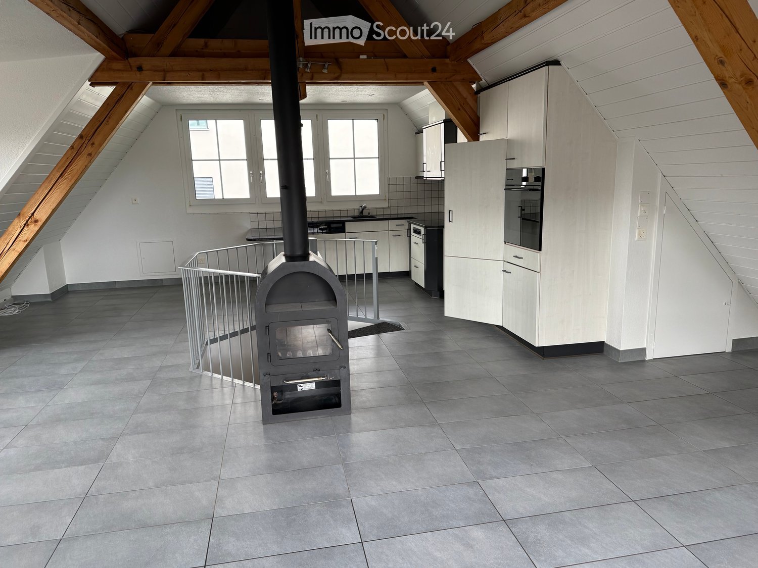 Loft-style room with exposed wooden beams, grey tiled floor, white kitchen cabinets, sink, faucet, oven, and a wood stove with a protective railing around it.