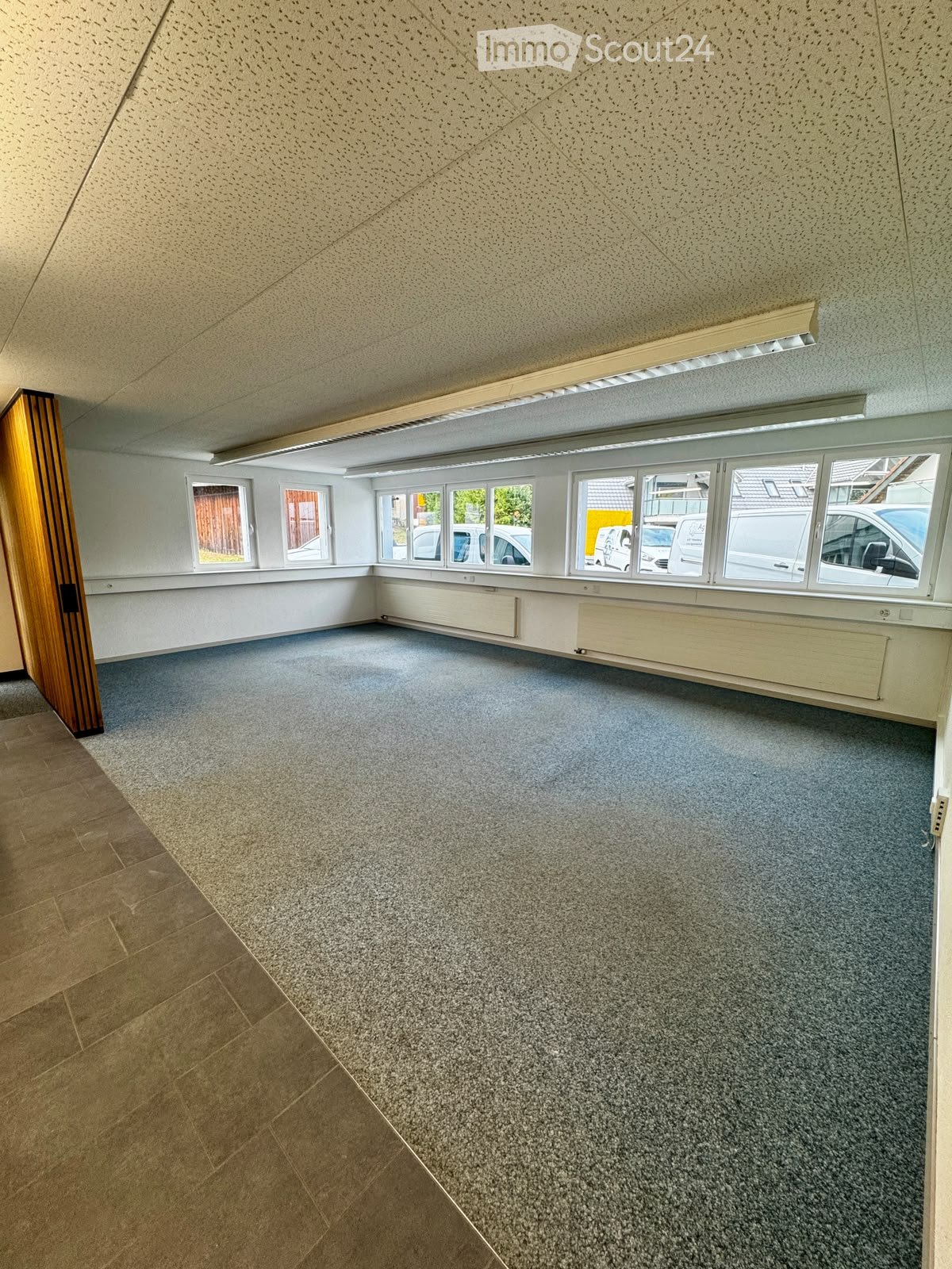 Empty room with grey carpeted floor, white walls, large windows, and a wooden door. No furniture or equipment present.