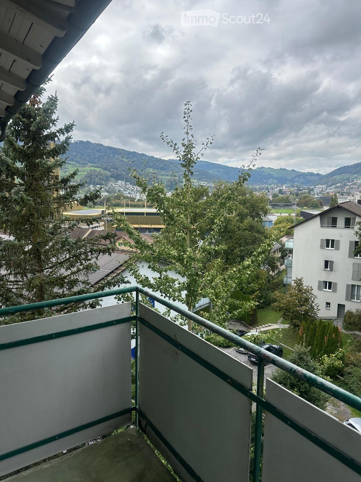 Balcony with metal railing overlooking a scenic view of a town, trees, and distant mountains.