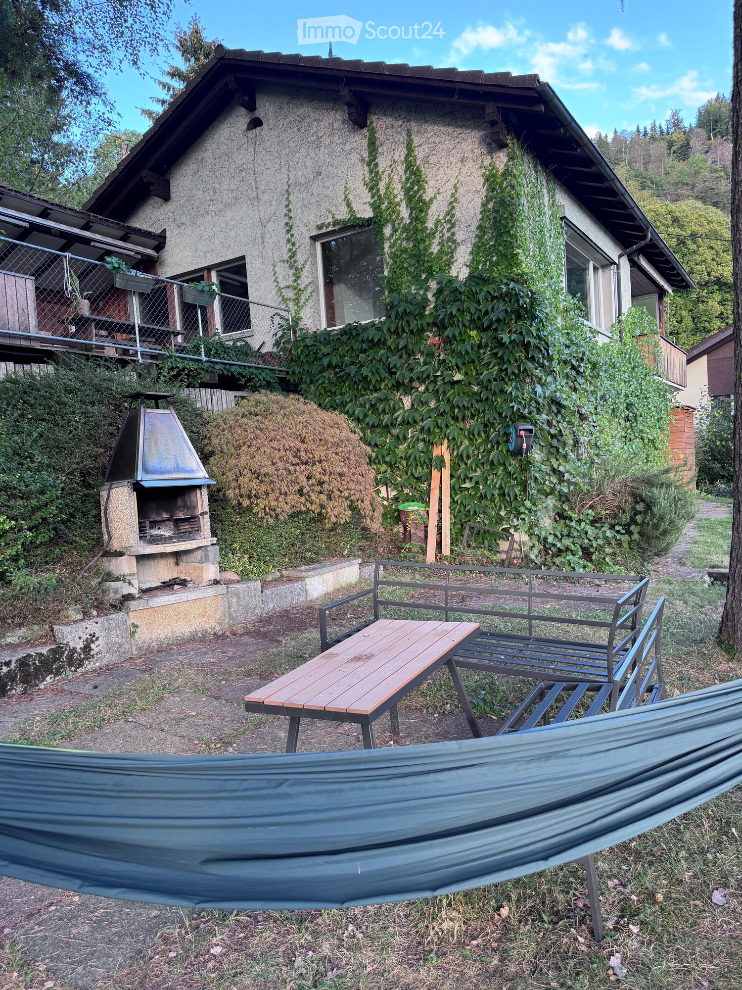 Outdoor area with a wooden table, metal chairs, a hammock, and a barbecue near a house covered in ivy.