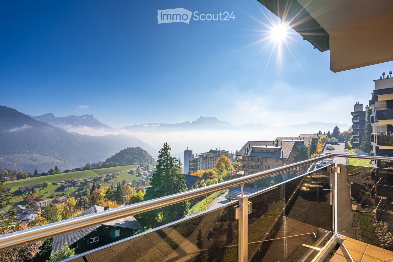 Balcony with a panoramic view of a town, surrounded by mountains and greenery, under a clear blue sky with the sun shining brightly.