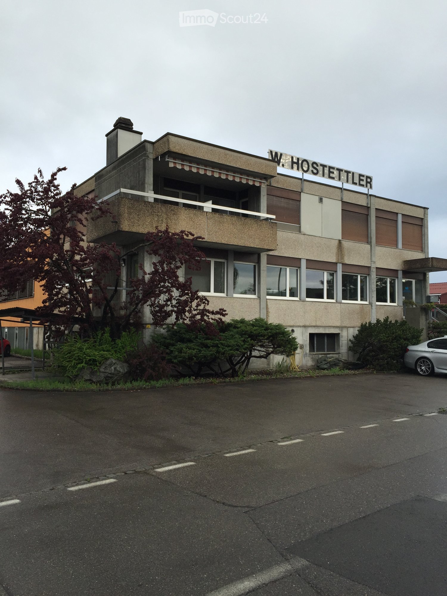 Two-story building with a balcony, signage reading "W. HOSTETTLER", surrounded by greenery and a parking area.