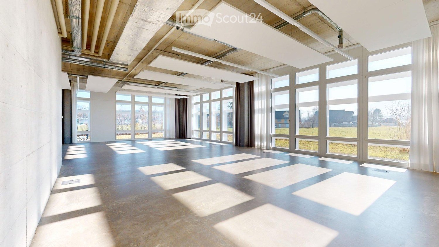 Empty room with large windows, exposed wooden beams, and industrial-style lighting fixtures. The floor is concrete, and the walls are white.