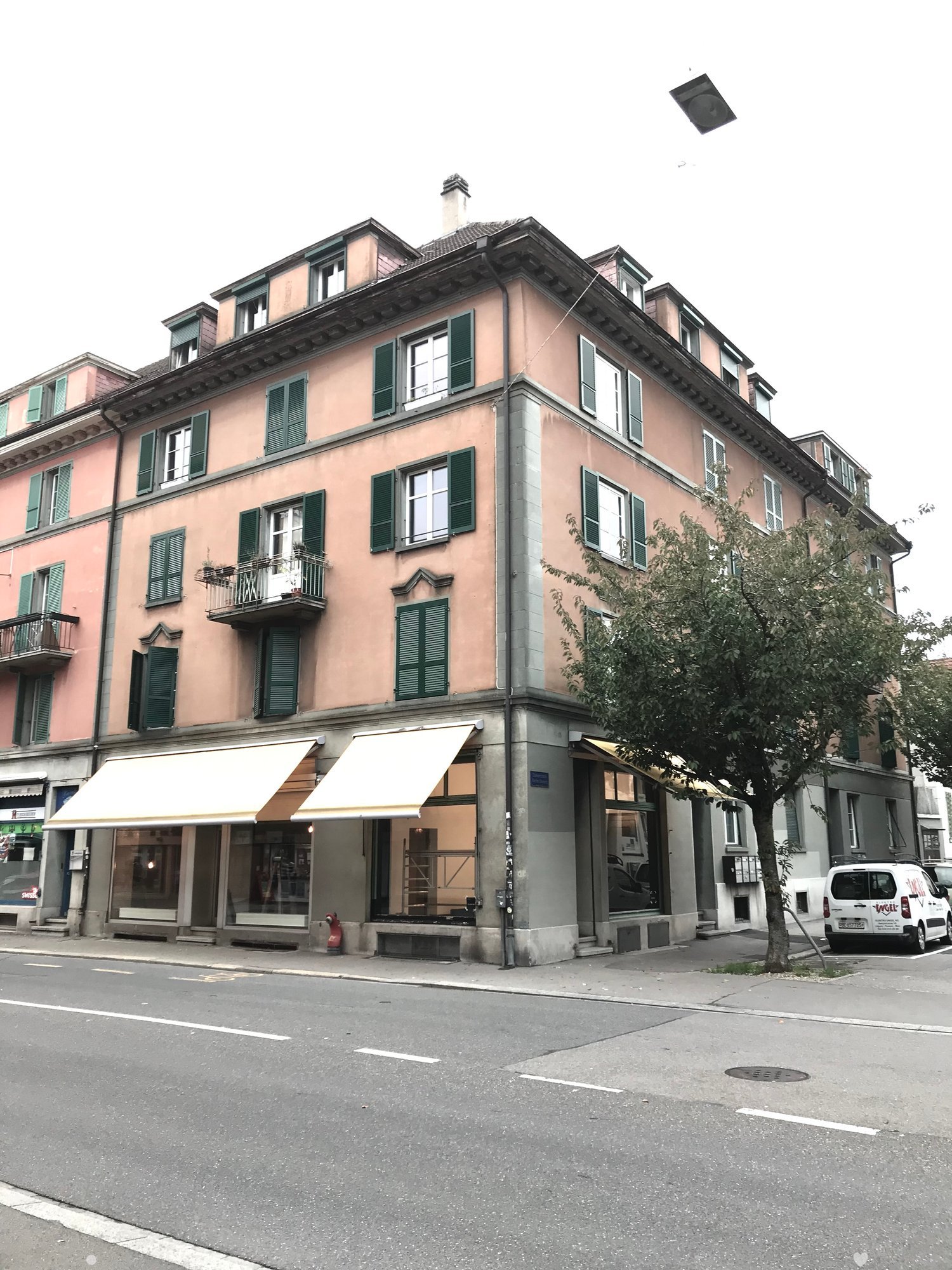 A four-story apartment building on a street corner with multiple windows and balconies on all levels. The facade is beige with large green shutters. There is an awning in front of the building, and a fire extinguisher is placed next to it. There are two t