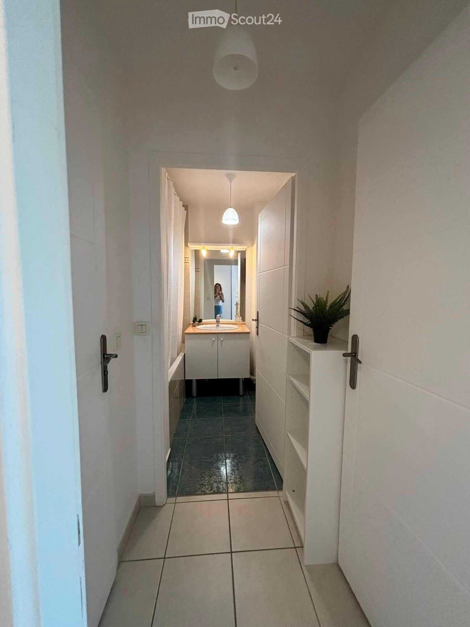 A narrow hallway with tiled floors, white walls, and a small bathroom visible at the end. The bathroom features a sink, mirror, and a small potted plant on a shelf.