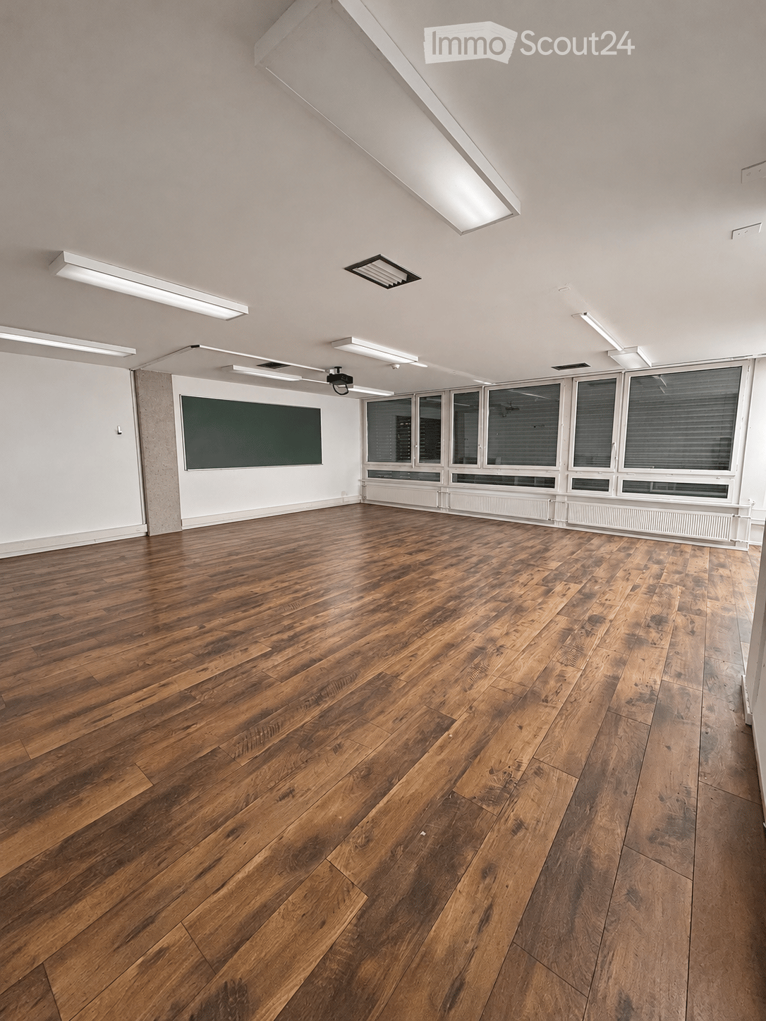 Empty room with wooden flooring, white walls, large windows, and a dark green chalkboard on one wall.