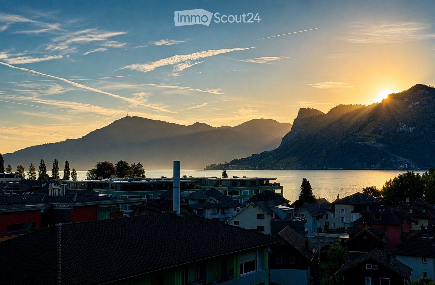 A scenic view of a town with colorful houses and rooftops, overlooking a calm lake and distant mountains during sunset.