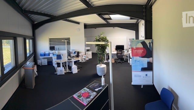 Modern office space with desks, chairs, computers, a potted plant, and a poster stand. The room features large windows and a high ceiling with exposed beams.