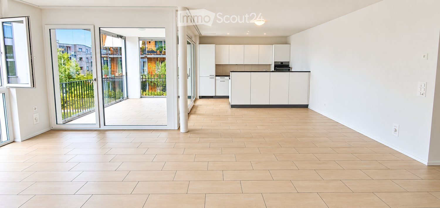 Bright, open-plan living and dining area with light wooden flooring, white kitchen cabinets, and a balcony with a metal railing.