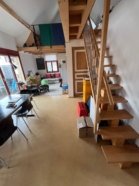 The image shows a modern, open-plan living and dining area with exposed wooden beams on the ceiling. The space features a wooden staircase leading to an upper level, a dining table with chairs, a sofa, a small ball, and a lamp. The room has large windows