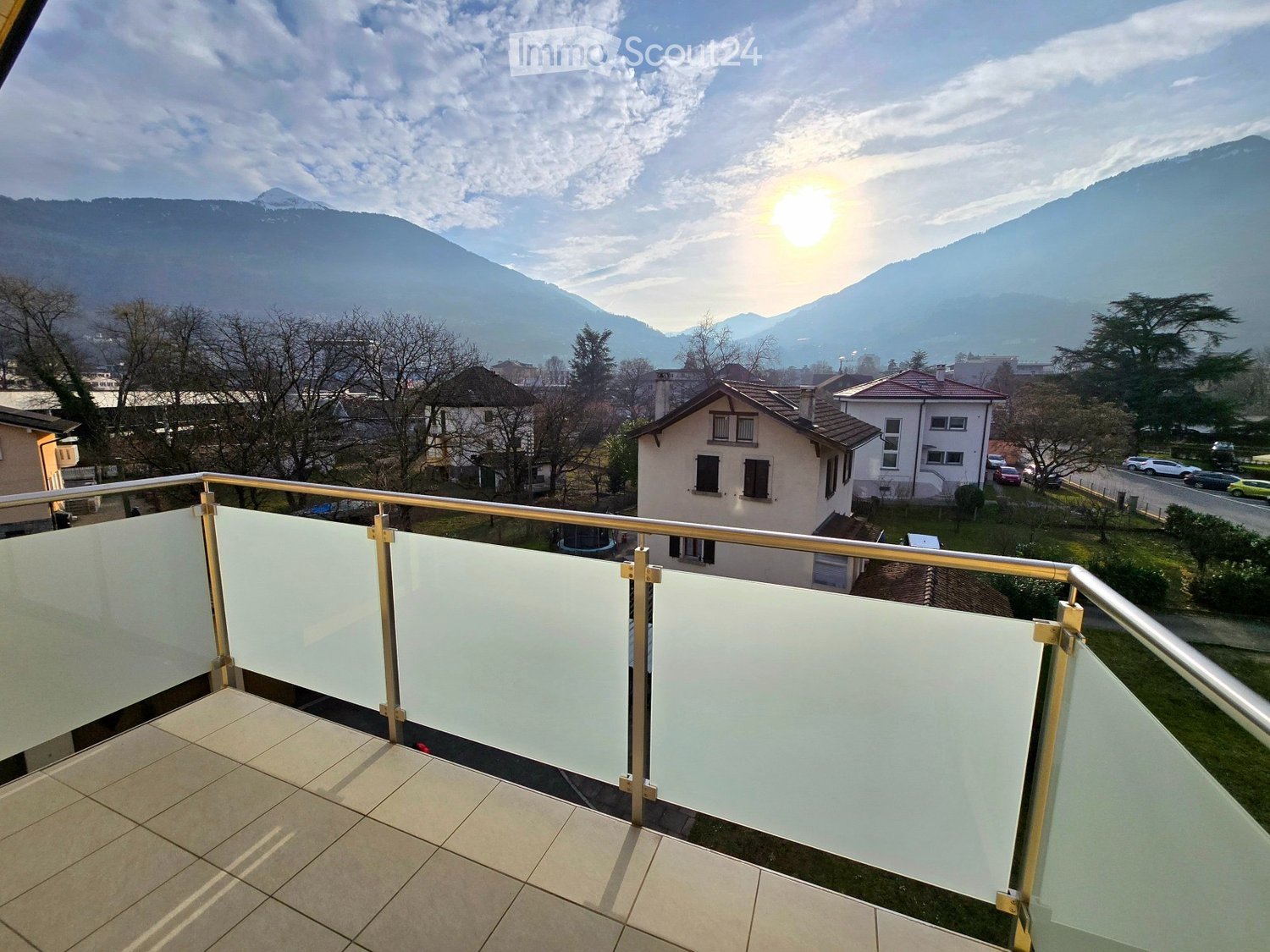 Balcony with glass railing, tiled floor, overlooking a town with houses and mountains in the background.