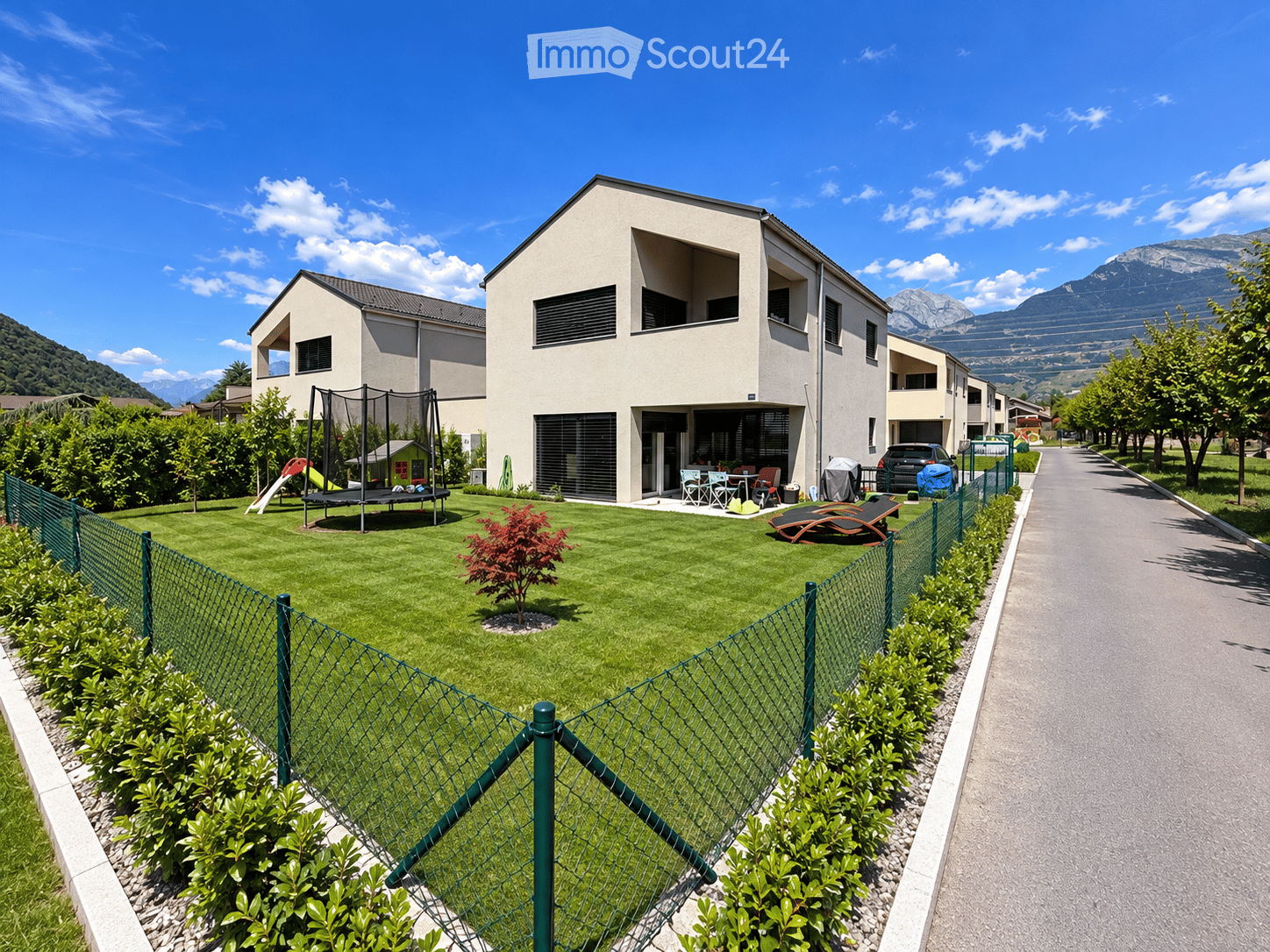 A modern residential complex with a well-maintained lawn, a trampoline, a slide, outdoor furniture, and lounge chairs, surrounded by a green fence and trees, with mountains in the background.