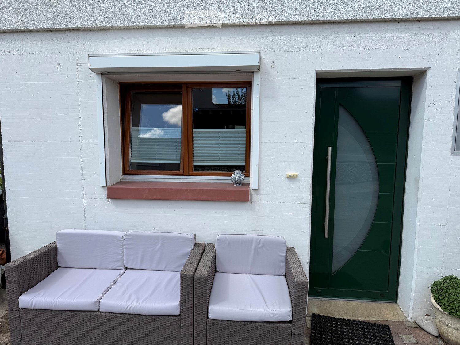 Outdoor seating area with a white cushioned sofa and chair, a small decorative vase on the windowsill, and a green front door.