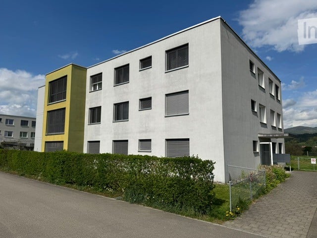 Modern multi-story building with white facade and grey accents, featuring rectangular windows and a neatly trimmed hedge along the front.
