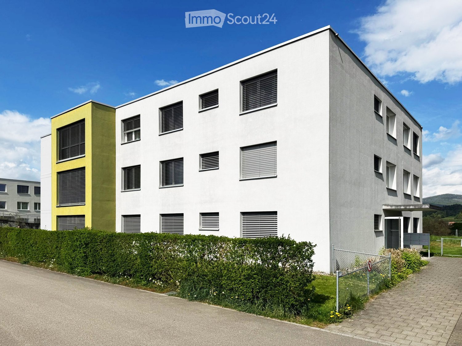 Modern multi-story building with white and yellow facade, multiple windows with blinds, surrounded by greenery and a paved driveway.