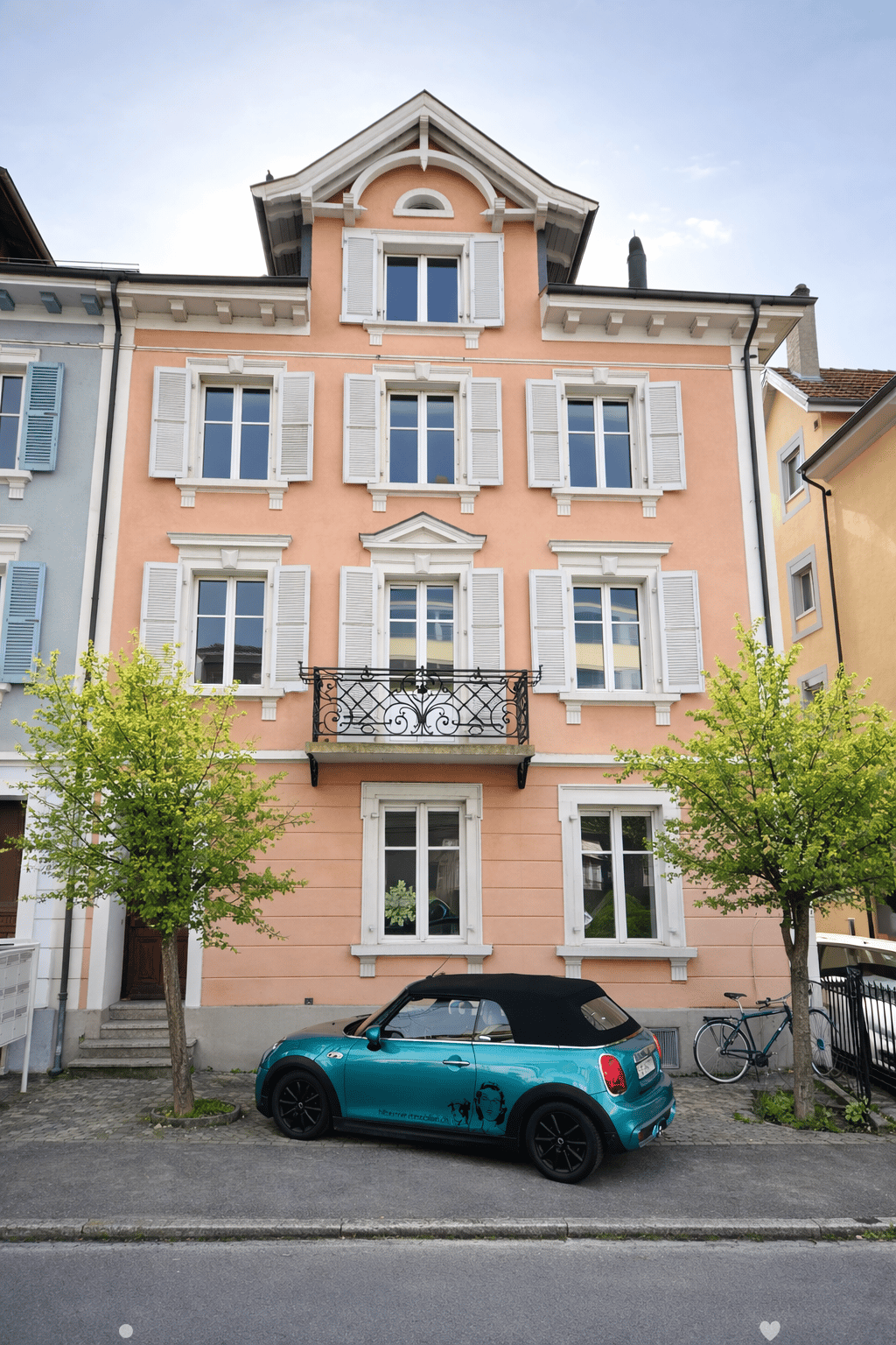 A two-story pink building with white window frames and shutters, featuring a balcony with a decorative railing. A turquoise convertible car is parked in front, and a bicycle is leaning against a fence.