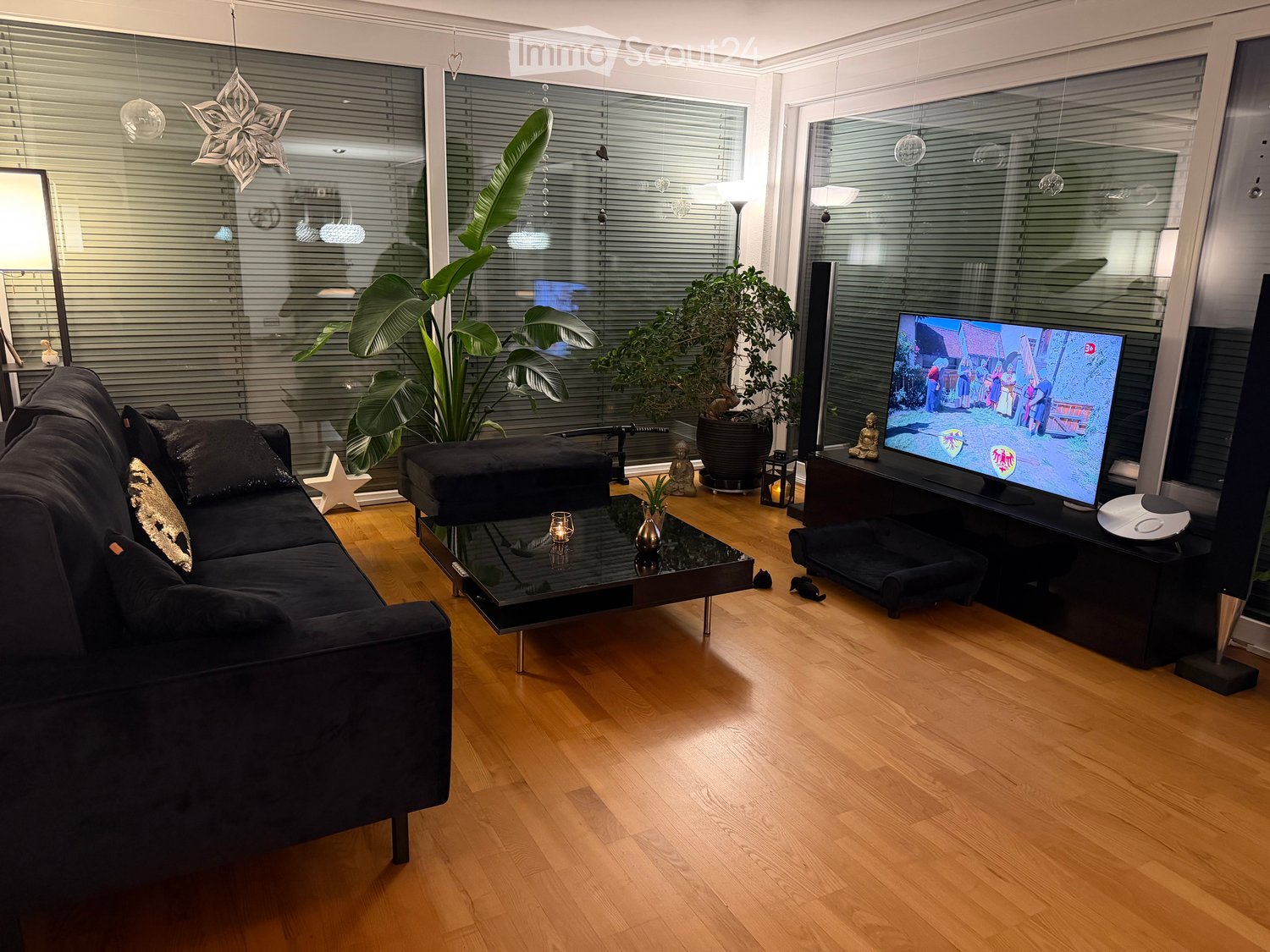 Modern living room with hardwood flooring, large windows, a black sectional sofa, coffee table, TV, and decorative plants.