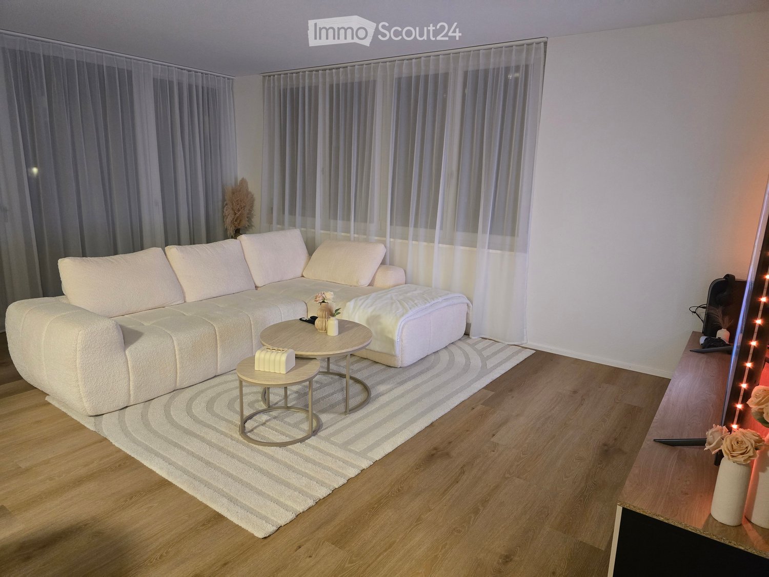 Modern living room with a white sectional sofa, ottoman, coffee table, small round table, and a television on a wooden stand. The room features a light-colored rug, wooden flooring, and sheer curtains.