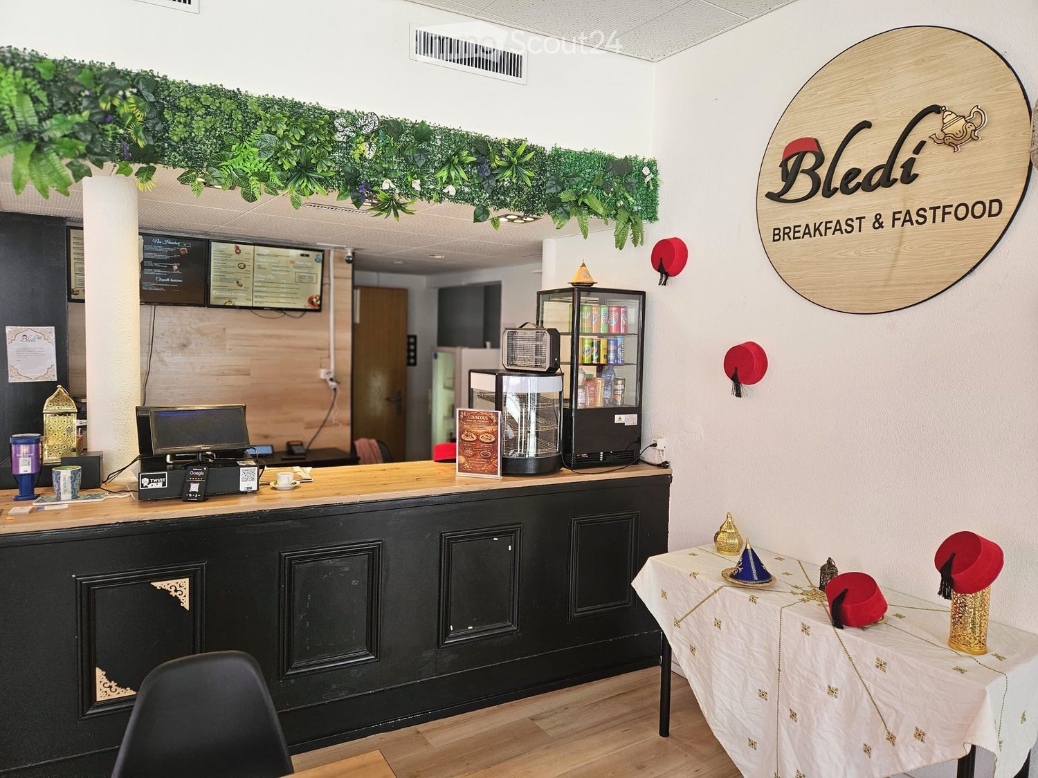 A modern breakfast and fast-food restaurant with a wooden counter, screens, a refrigerator, and a small table with decorative hats.