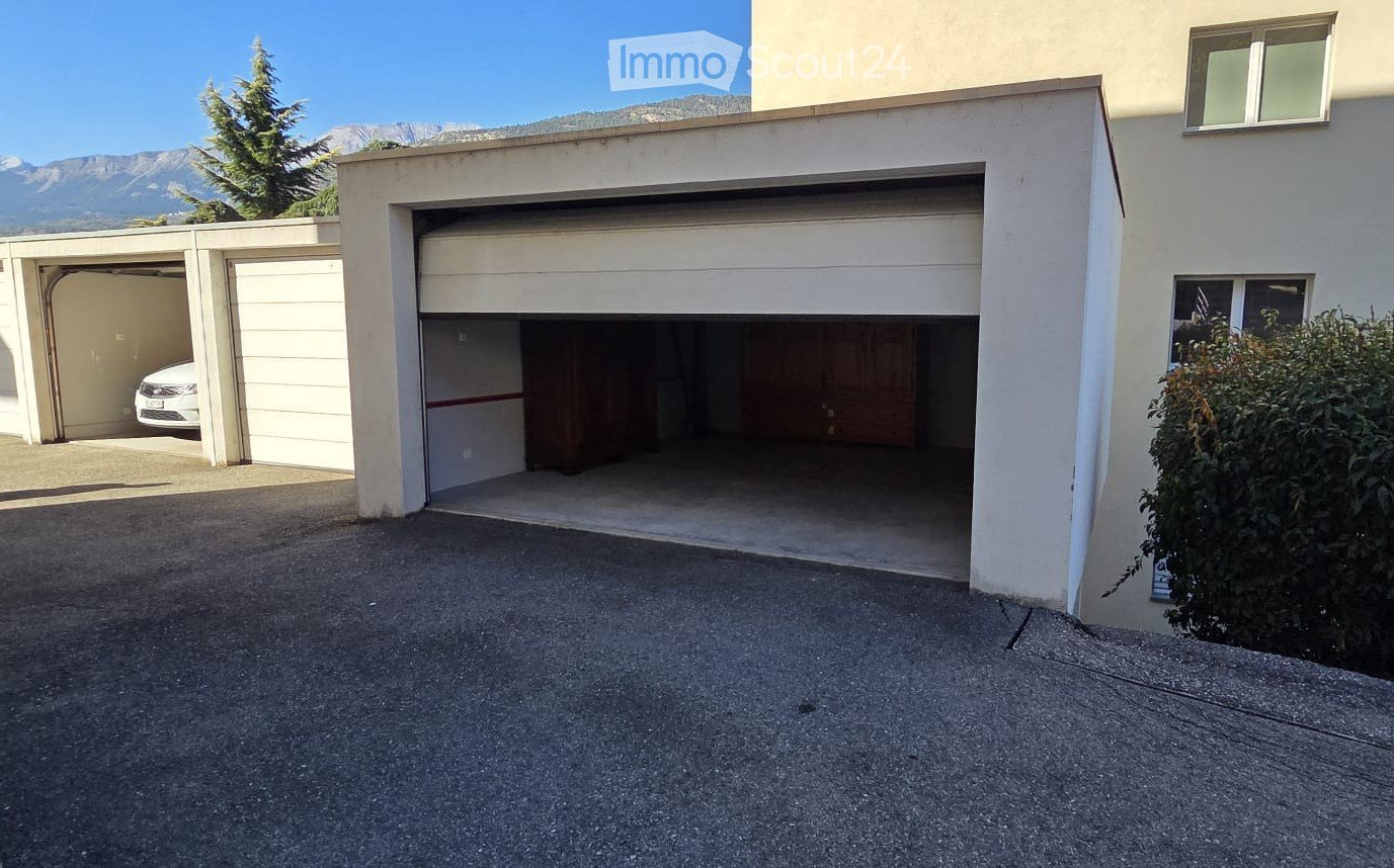 Two-car garage with a white car parked inside, located next to a building with a raised ground floor and a view of mountains in the background.