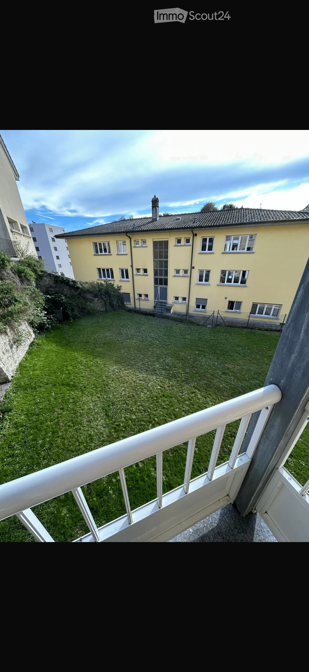 Balcony overlooking a grassy area with a yellow building in the background, featuring white railings and a view of the sky.