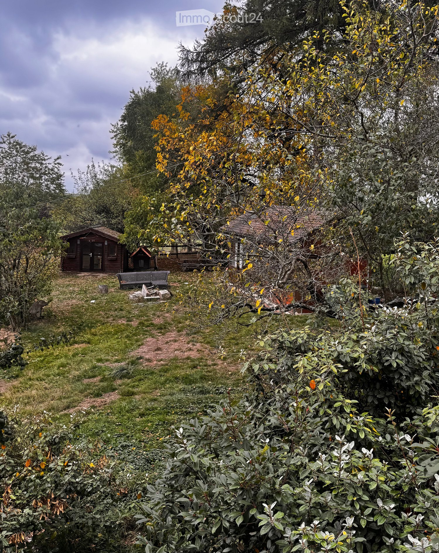 A serene garden area with a wooden shed, benches, and lush greenery, including trees with yellow leaves and a variety of shrubs and plants.