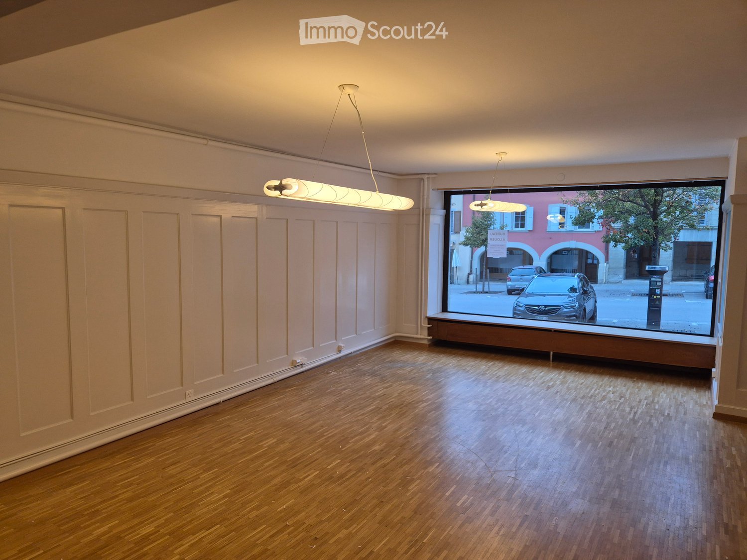 Empty room with wooden flooring, light-colored walls, and large windows showing a street view. No furniture or equipment present.