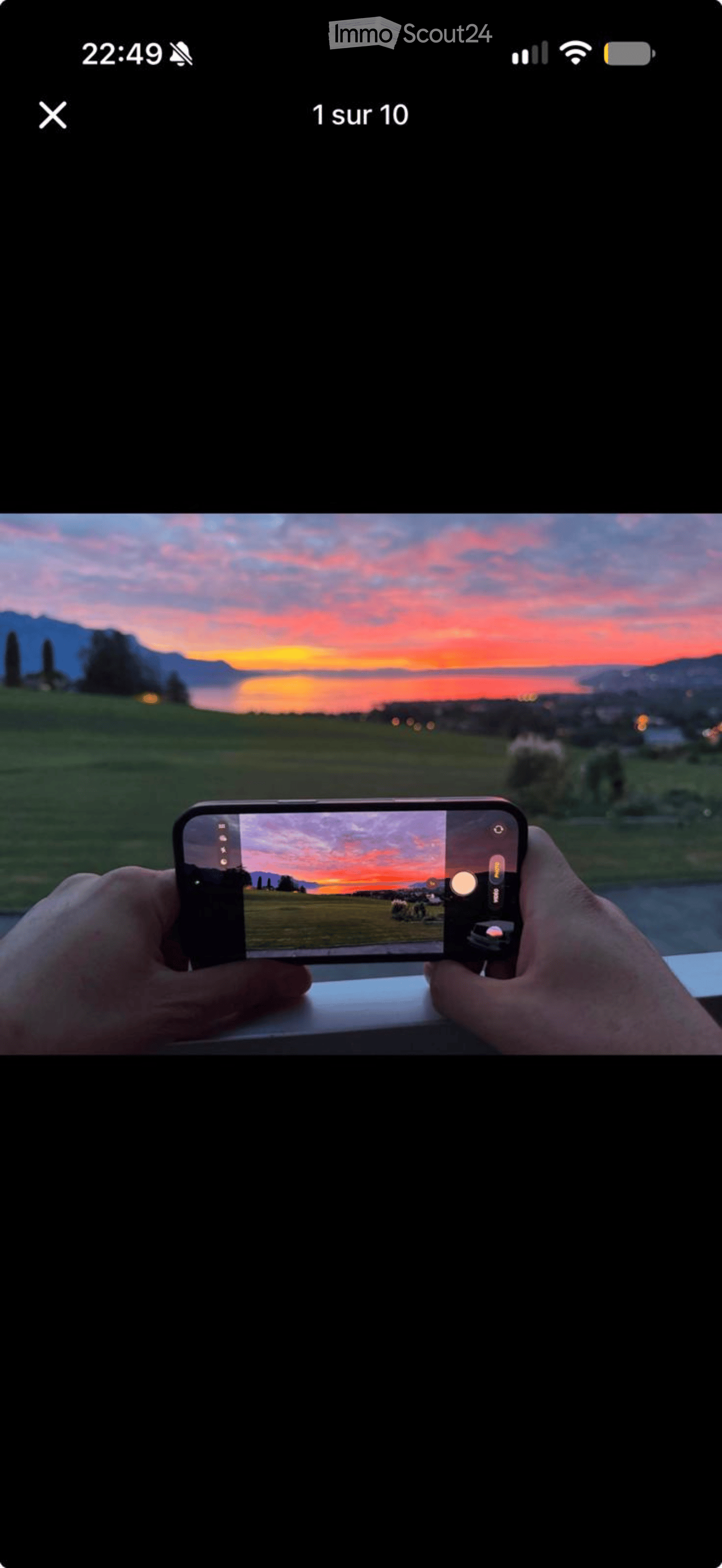 A person is taking a photo of a scenic sunset view over a lake and distant mountains using a smartphone.