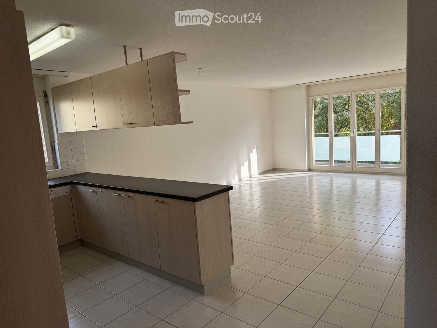 Empty kitchen with light wood cabinets, black countertop, tiled floor, and large windows leading to a balcony.