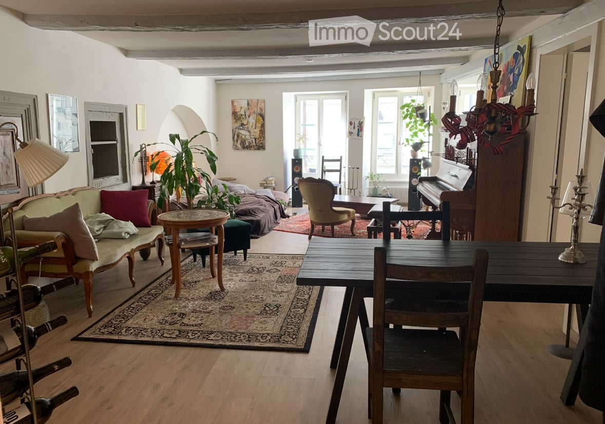 A spacious, well-lit living and dining area with wooden floors, a patterned rug, a sofa, a dining table, chairs, a piano, and decorative lamps.