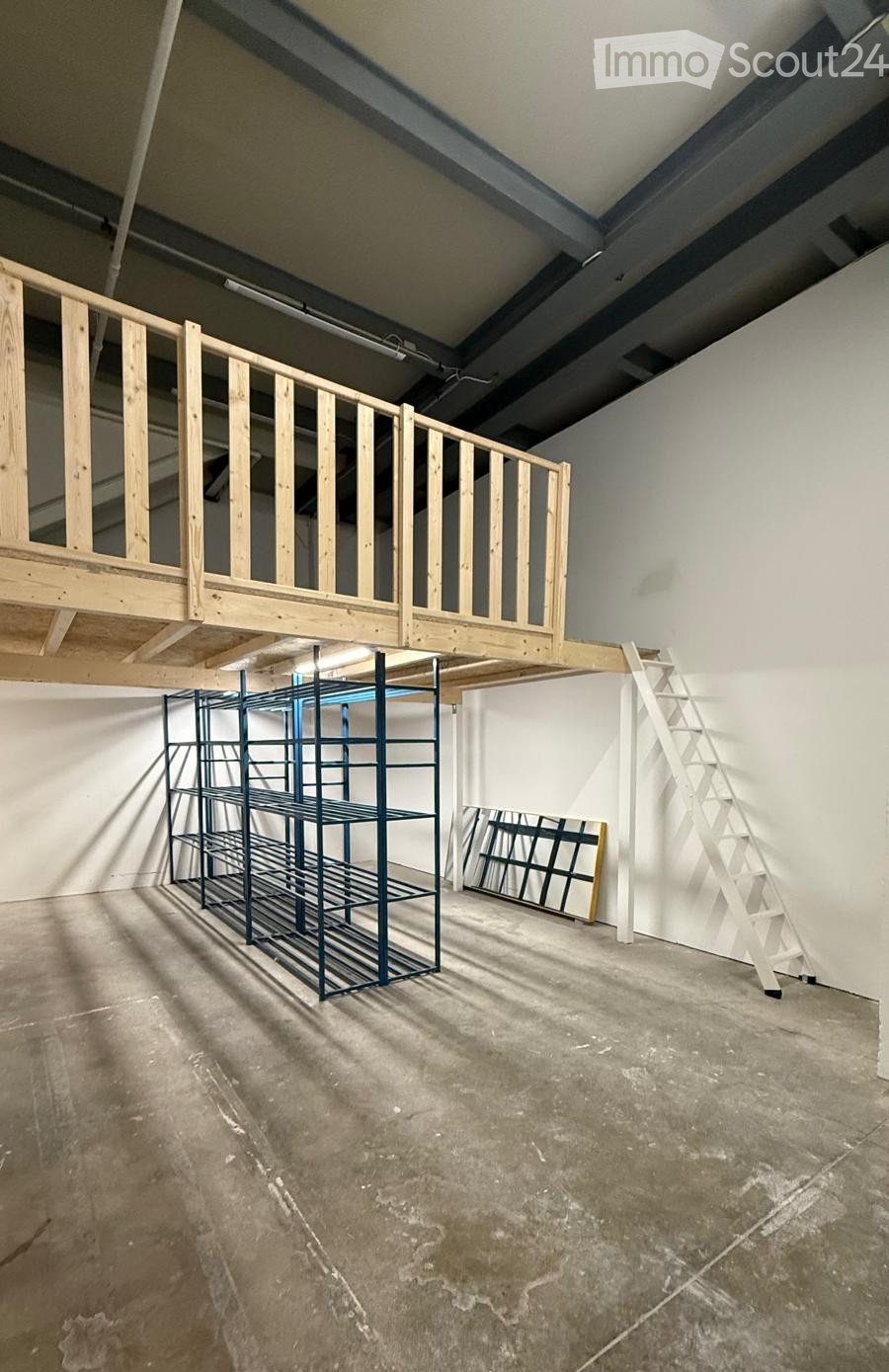 Empty room with unfinished concrete floor, white walls, exposed metal beams, and a wooden loft platform with a ladder leading up to it. A metal shelving unit and a large mirror are present.