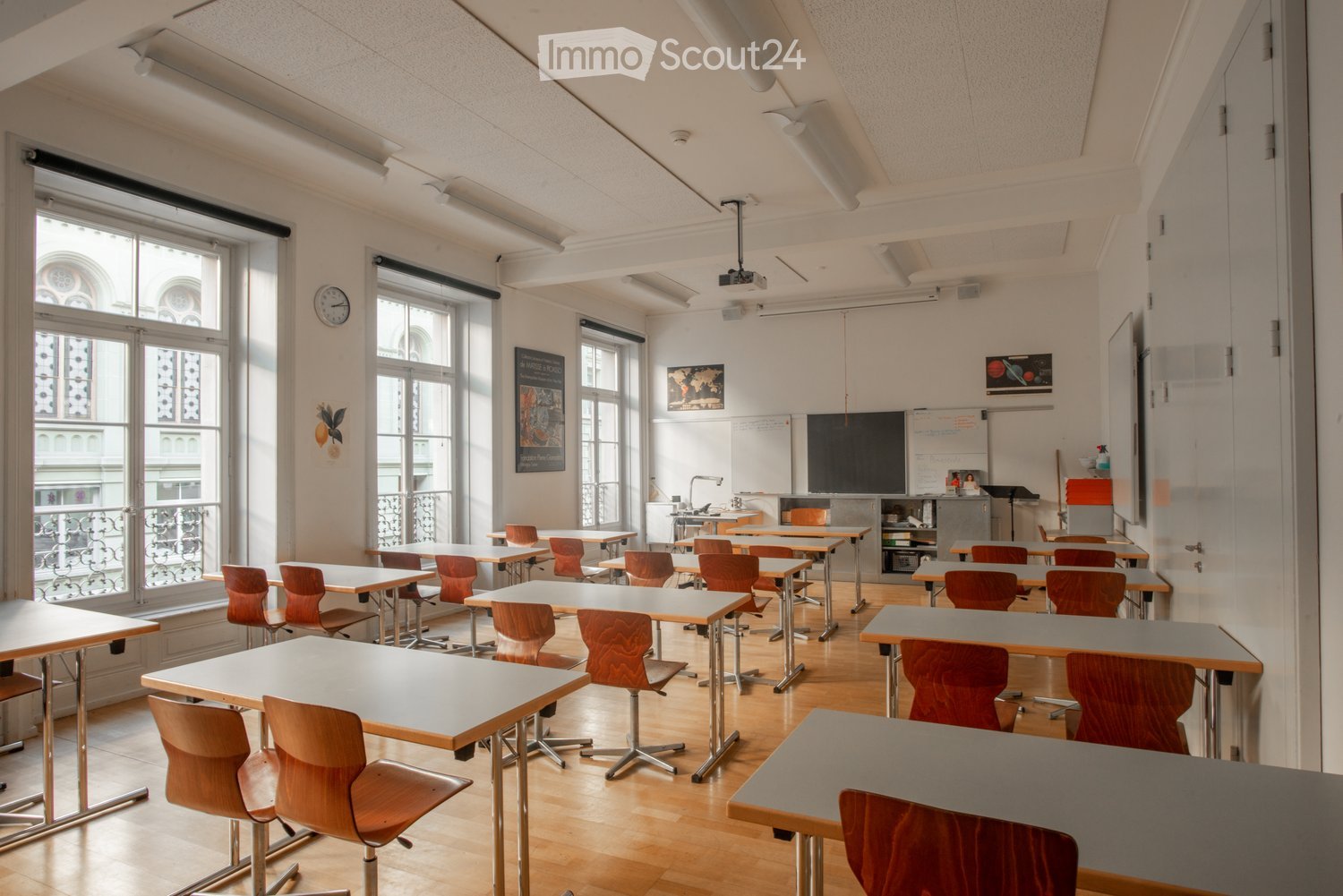 Bright classroom with wooden tables and chairs, large windows, a projector screen, and educational posters on the walls.