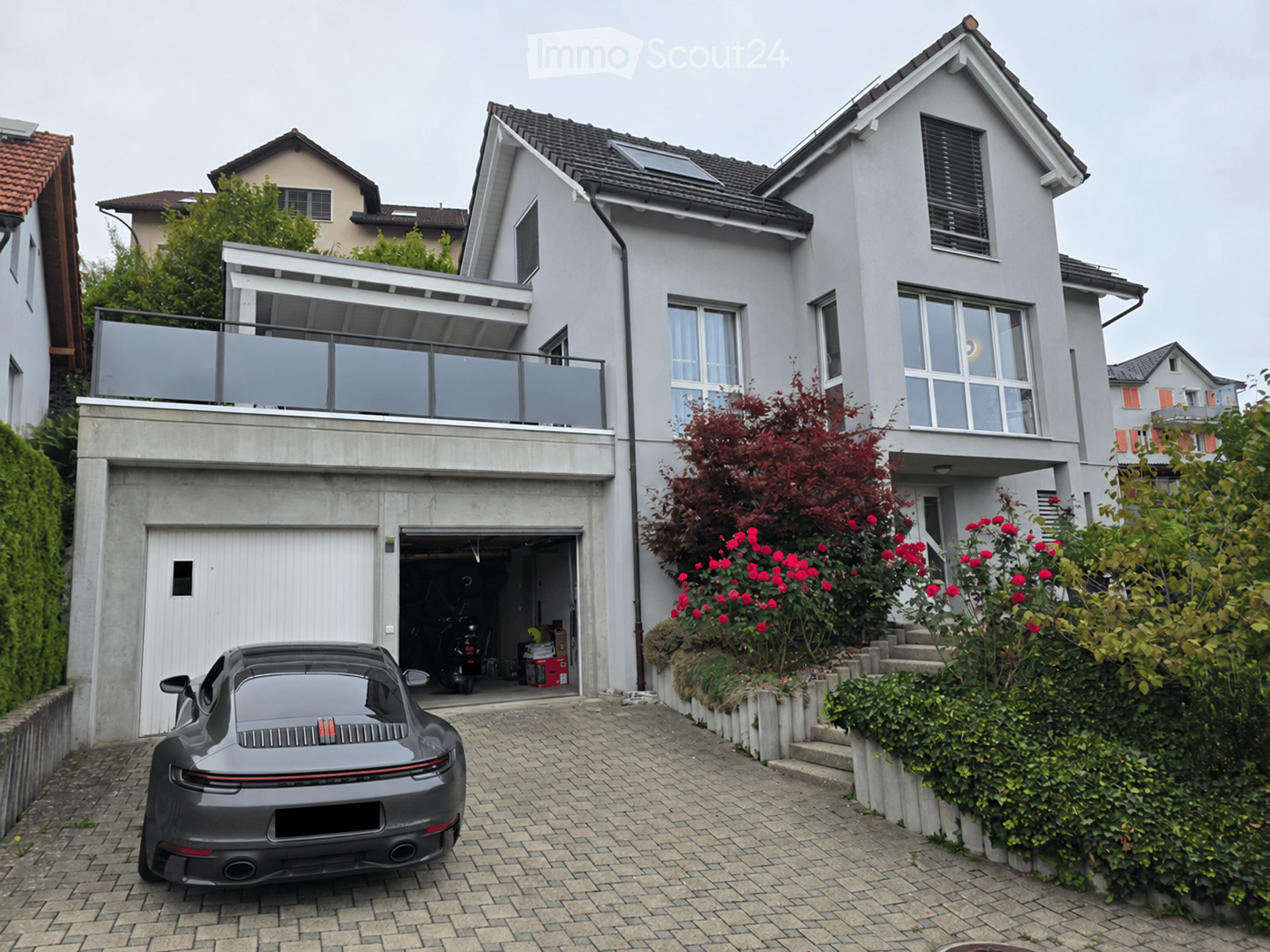 Modern two-story house with grey exterior, large windows, balcony, garage, and well-maintained garden with red flowers.