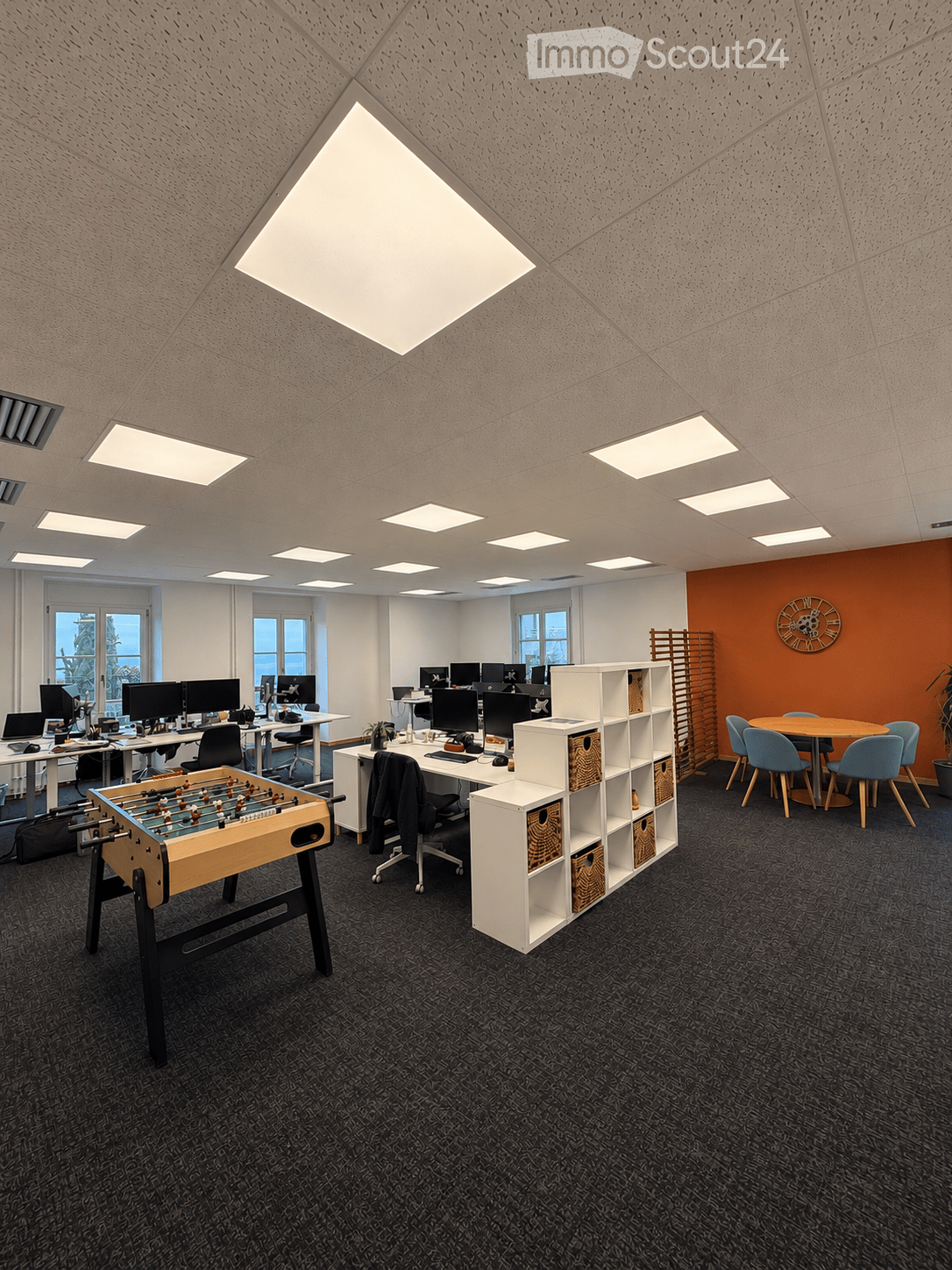 Modern office space with multiple workstations, a foosball table, and a round meeting table with chairs.